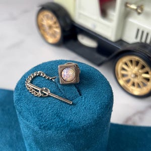 May include: A silver tie tack featuring a small, iridescent, square stone. The tie tack is connected to a chain and a small bar. It's displayed on a teal velvet stand, with a toy car in the background.