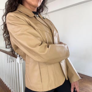 May include: A tan leather jacket with a collared neckline and two decorative buttons. The jacket has two front pockets and a tailored fit. The sleeves are long and the jacket appears to be made of a soft leather material.