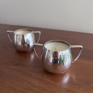 Może przedstawiać: Dwa srebrne świeczniki o okrągłym, lekko zakrzywionym kształcie. Każdy świecznik ma dwa uchwyty i zawiera białą świecę.