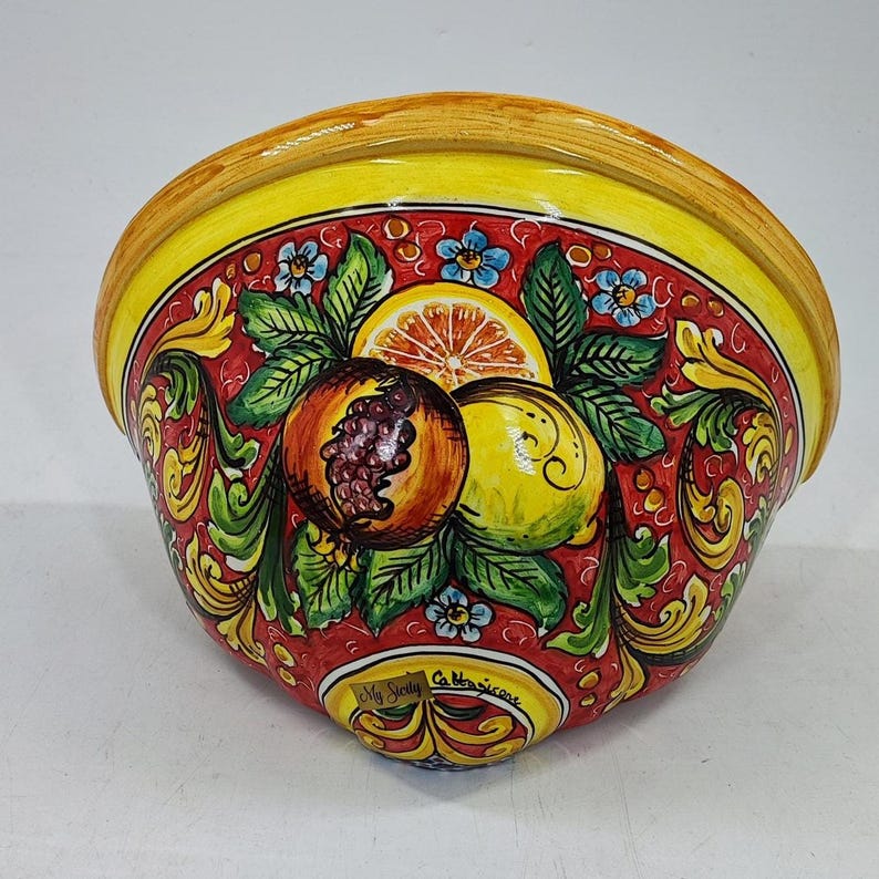 May include: A ceramic wall pocket with a vibrant design. The bowl features a red background with painted fruit, including a pomegranate, lemon, and orange. The rim is yellow, and the design includes green leaves and blue flowers. The text "My Sicily" is visible.
