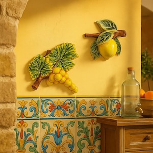 Può includere: Decorazioni murali in ceramica raffiguranti un limone e un grappolo d'uva, ciascuno con foglie verdi e steli marroni. L'arte è posta su una parete gialla, sopra piastrelle decorate. Sono visibili anche una bottiglia di vetro e un mobile in legno.