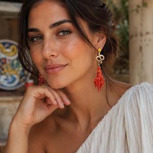 Könnte beinhalten: Eine Frau trägt goldene Schlangenohrringe mit roten, korallenartigen Anhängern. Die Ohrringe bilden einen auffälligen Kontrast zu ihrer hellen Haut und dem weißen One-Shoulder-Top. Die Ohrringe verleihen dem Gesamtbild einen Hauch von Eleganz und Farbe.