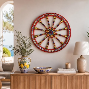 Può includere: Una grande ruota di carro decorativa in legno rosso con intricati motivi floreali e geometrici dipinti a mano in blu, giallo e verde, dotata di un mozzo centrale e dodici raggi che si irradiano verso l'esterno fino al cerchio.