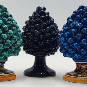 Puede incluir: Tres esculturas de cerámica de piñas en verde azulado, negro y azul. Cada una tiene una superficie detallada y texturizada y se asienta sobre una base decorativa con un diseño estampado. Las bases son naranjas.