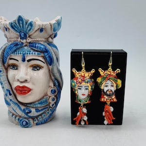 Könnte beinhalten: Eine Keramikvase und ein Paar Ohrringe. Die Vase ist weiß und blau mit einem Gesichtsdesign. Die Ohrringe haben goldene Haken, Gesichtsanhänger, Korallen und Perlenakzente. Die Ohrringe werden auf einer schwarzen Box präsentiert.