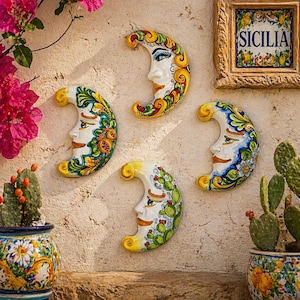 Peut inclure: Quatre décorations murales en forme de croissant de lune en céramique, avec des visages peints et des motifs floraux et botaniques colorés. Les lunes sont blanches avec des accents jaunes, verts, bleus et rouges. Une tuile encadrée avec le mot "SICILIA" est également visible.