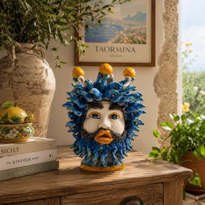 Peut inclure: Un vase en céramique en forme de visage avec une barbe et une couronne bleues. Le visage est blanc avec des yeux bleus et une moustache orange. Le vase est sur une surface en bois avec des livres et un petit pot à citron décoratif.