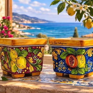 Könnte beinhalten: Zwei quadratische Keramik-Pflanzgefäße mit handgemalten Designs. Eines zeigt Zitronen und das andere Granatäpfel, beide mit gelben und blauen Akzenten. Die Pflanzgefäße haben einen gelben Rand und sind vor einer Küstenkulisse platziert.