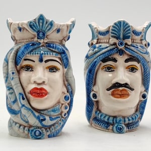 Könnte beinhalten: Zwei keramische Vasen in Kopfform. Eine zeigt eine Frau mit roten Lippen und blauen Akzenten, die andere einen Mann mit Schnurrbart. Beide haben verzierten Kopfschmuck und sind in Weiß, Blau und Beige bemalt.