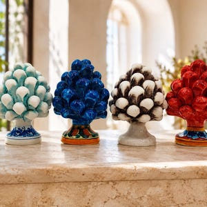 May include: Four ceramic pinecone sculptures arranged in a row. The first is light blue and white, the second is deep blue with a patterned base, the third is white with dark grey accents, and the fourth is bright red with a colourful base.