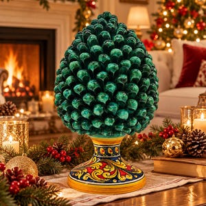 Puede incluir: Un adorno de piña de cerámica con un acabado verde oscuro brillante y un patrón de escamas texturizado. La base está decorada con motivos florales pintados a mano en amarillo, rojo y azul sobre fondo blanco, sobre un pedestal circular.