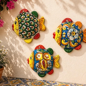 May include: Three hand-painted ceramic fish wall plaques featuring vibrant, multi-colored patterns. Each fish has a distinct design, including green scales, floral motifs, and fruit illustrations, with yellow faces, red lips, and integrated hanging holes.