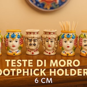May include: Six ceramic Testa di Moro toothpick holders, each 6 cm tall, with detailed faces and colorful accents. The holders are arranged on a wooden surface, showcasing their unique design.