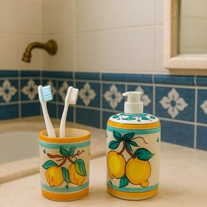 Puede incluir: Un juego de baño de cerámica con un porta cepillos de dientes con dos cepillos y un dispensador de jabón. El juego está decorado con un diseño de limón en amarillo, verde y azul, sobre un fondo blanco con un borde amarillo y azul.
