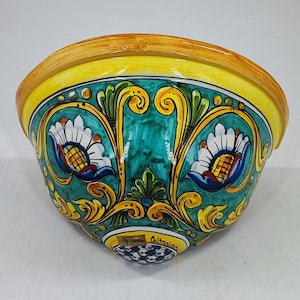 May include: A hand-painted ceramic wall pocket with a vibrant design. The bowl features a yellow rim, teal interior, and intricate floral patterns in white, blue, and yellow. The base has a small, decorative pattern and the text "My Sicily Caltagirone".