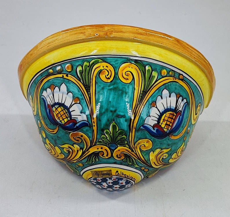 May include: A hand-painted ceramic wall pocket with a vibrant design. The bowl features a yellow rim, teal interior, and intricate floral patterns in white, blue, and yellow. The base has a small, decorative pattern and the text "My Sicily Caltagirone".