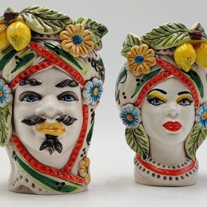 Puede incluir: Dos jarrones de cerámica con forma de cabeza, uno masculino y otro femenino, con detalles intrincados. El jarrón masculino tiene bigote y barba, mientras que el femenino tiene labios rojos y sombra de ojos amarilla. Ambos están adornados con limones, flores y hojas verdes.