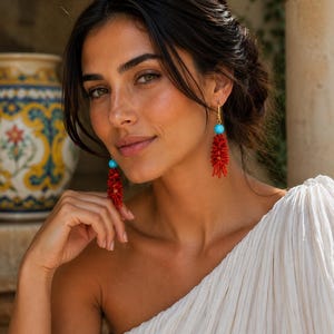 May include: A woman wearing red coral earrings with turquoise beads. The earrings are suspended from gold hooks. She is wearing a white, one-shoulder dress. The background is a blurred outdoor scene with stone columns.