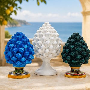 Puede incluir: Tres esculturas de cerámica con forma de piña dispuestas en fila. La escultura de la izquierda es azul brillante, la del centro es blanca sólida y la de la derecha es verde oscuro. Las bases de las piezas azul y verde tienen motivos florales.