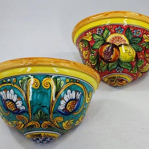 May include: Two ceramic bowls with vibrant hand-painted designs. One bowl features a red background with fruit and floral motifs, while the other has a teal background with floral patterns. Both have yellow rims.