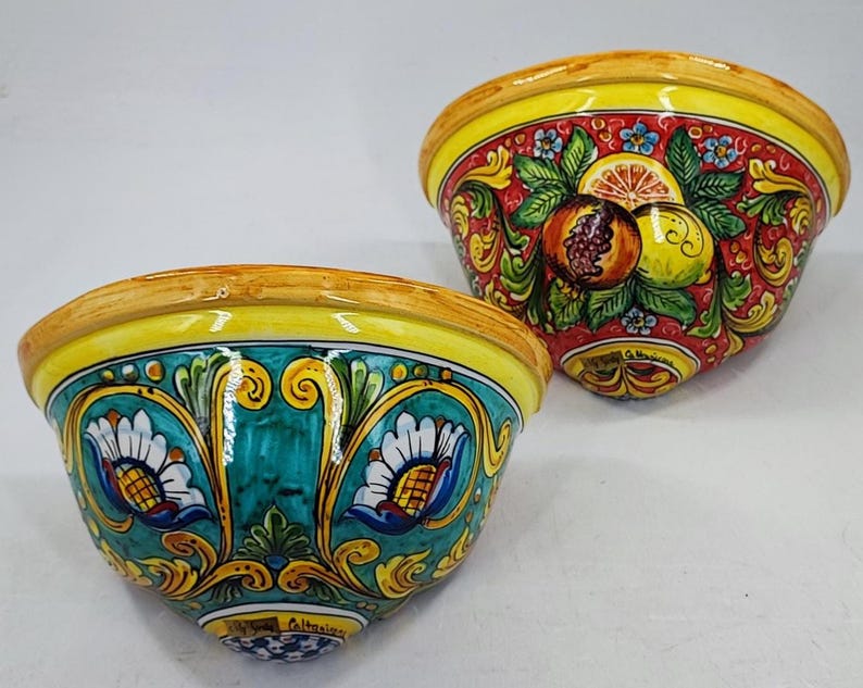 May include: Two ceramic bowls with vibrant hand-painted designs. One bowl features a red background with fruit and floral motifs, while the other has a teal background with floral patterns. Both have yellow rims.