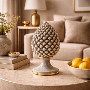 May include: A ceramic pinecone ornament with a grey and gold base. The pinecone is light beige with detailed texture. The ornament sits on a round, marble-effect table, with a bowl of lemons and a vase of greenery in the background.