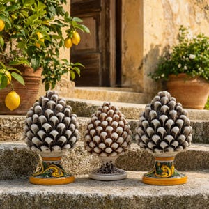 Op de afbeelding: Drie keramische dennenappelsculpturen op stenen treden. De twee buitenste stukken hebben witte en donkergrijze schubben met gele en groene decoratieve voetstukken. Het middelste stuk heeft witte en bruine schubben met een witte voet.