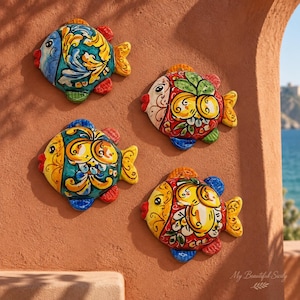 Op de afbeelding: Vier keramische vis-wanddecoraties gerangschikt op een getextureerd terracotta oppervlak. Twee vissen hebben blauwe en gele patronen, terwijl de andere twee rode en gele ontwerpen tonen. Elke vis heeft een rond lichaam en rode lippen.