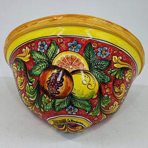 May include: A ceramic bowl with a vibrant design. The bowl features a yellow rim and a red interior adorned with painted fruits, green leaves, and blue flowers. The fruits include a pomegranate, lemon, and orange slice. The text "My Sicily Caltagirone" is visible.