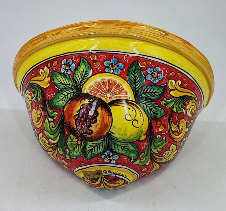 May include: A ceramic bowl with a vibrant design. The bowl features a yellow rim and a red interior adorned with painted fruits, green leaves, and blue flowers. The fruits include a pomegranate, lemon, and orange slice. The text "My Sicily Caltagirone" is visible.