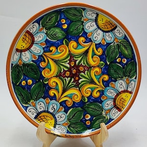 Può includere: Un piatto in ceramica dipinto a mano con un motivo floreale. Il piatto presenta uno sfondo blu scuro con fiori bianchi, gialli e blu, foglie verdi e un bordo marrone. Il piatto è esposto su un supporto di legno.
