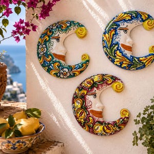Puede incluir: Tres placas de pared en forma de media luna hechas de cerámica, cada una con un rostro pintado a mano con distintos patrones florales en azul, verde y rojo. Las lunas tienen un acabado brillante y están expuestas sobre una pared blanca.
