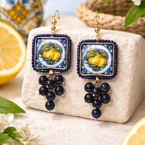May include: Gold-toned earrings featuring square ceramic tiles with lemon illustrations, framed in blue beads. Dark blue bead clusters dangle below. The earrings are displayed on a stone surface, with a lemon half visible.