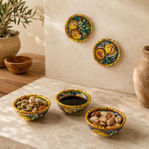 Könnte beinhalten: Fünf kleine, runde Keramikschalen mit handbemalten gelben, grünen und blauen Fruchtmotiven. Zwei Schalen sind an der Wand befestigt, während drei auf einer flachen Oberfläche stehen, wobei zwei Nüsse und eine eine dunkle Flüssigkeit enthalten.