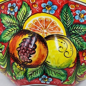 May include: A hand-painted ceramic bowl with a vibrant design. The bowl features a yellow rim and a red interior adorned with illustrations of a pomegranate, lemon, and orange slice. Green leaves and blue flowers complete the design. The text "My Sicily Cattasicare" is visible.