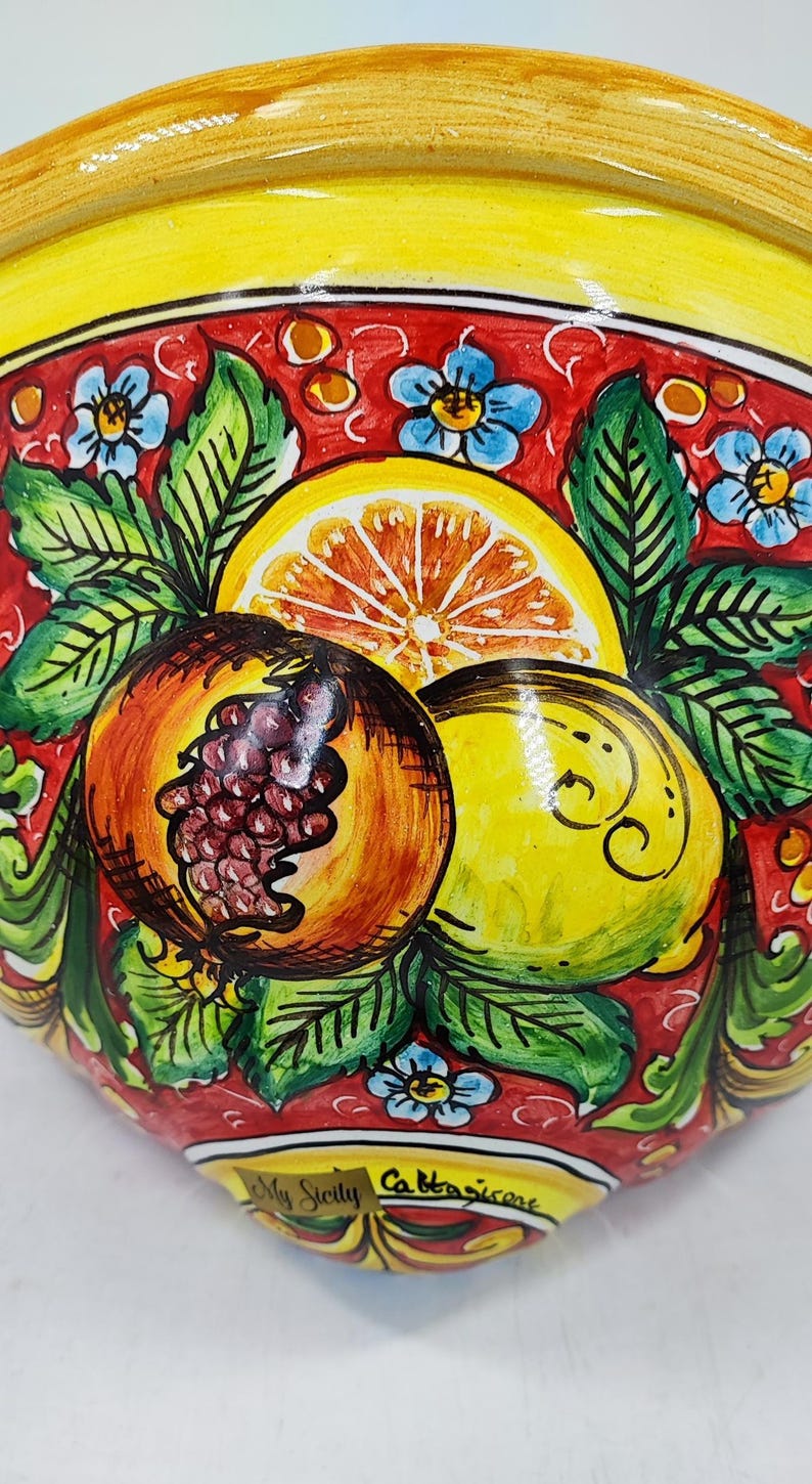 May include: A hand-painted ceramic bowl with a vibrant design. The bowl features a yellow rim and a red interior adorned with illustrations of a pomegranate, lemon, and orange slice. Green leaves and blue flowers complete the design. The text "My Sicily Cattasicare" is visible.