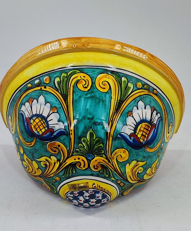 May include: A ceramic wall pocket with a vibrant, hand-painted design. The bowl features a yellow rim, teal interior, and ornate floral patterns in yellow, white, and blue. The base has a decorative pattern and text that reads "My Sicily Caltagirone."