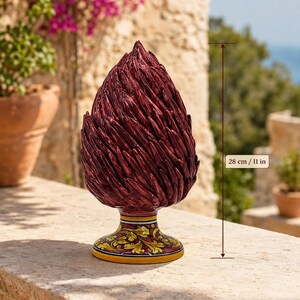 May include: A dark red ceramic pinecone sculpture with a textured, layered surface. The base features a yellow and green floral pattern. A vertical measurement line indicates a total height of 28 cm. The object stands on a stone ledge.