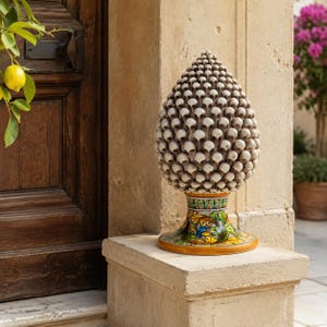 Könnte beinhalten: Eine Keramikskulptur in Form eines Tannenzapfens mit einer strukturierten, cremefarbenen Oberfläche aus sich wiederholenden schuppenartigen Mustern. Der Fuß ist mit handgemalten floralen Motiven in Grün, Gelb, Blau und Orange verziert.