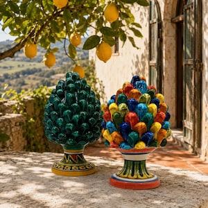 Puede incluir: Dos esculturas de cerámica en forma de piña sobre una superficie de piedra. Una es de color verde oscuro sólido con una base estampada en amarillo y azul. La otra es multicolor con escamas amarillas, azules, rojas y verdes sobre una base a rayas.