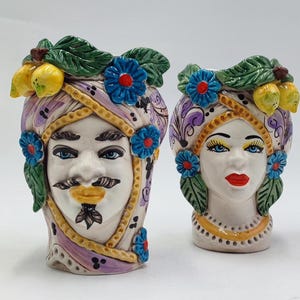 Puede incluir: Un par de jarrones de cerámica con forma de rostro humano. Los jarrones están decorados con flores, hojas y limones de colores. Los rostros están pintados con rasgos realistas, incluidos ojos, nariz, boca y cejas.