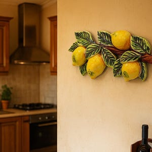 Può includere: Decorazione murale a forma di ramo di limone in ceramica con limoni giallo brillante e foglie verdi. I limoni sono attaccati a un ramo marrone. La decorazione è montata su una parete beige in una cucina.