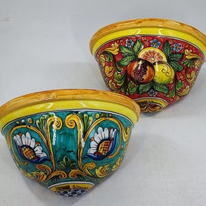 May include: Two decorative ceramic bowls. One bowl features a red background with fruit and floral designs. The other bowl has a teal background with floral designs. Both bowls have yellow rims.
