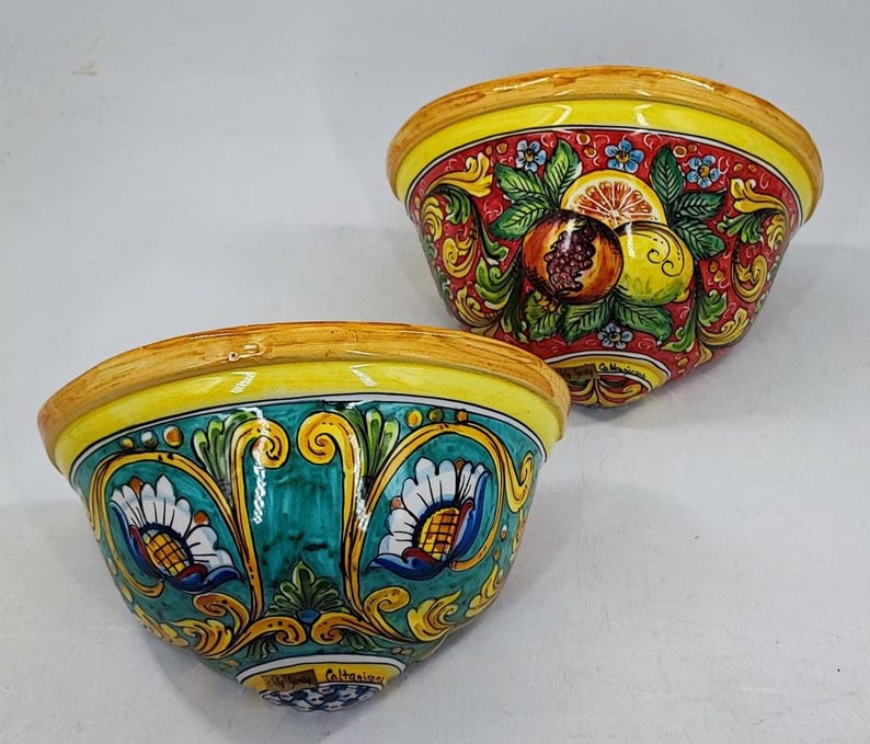 May include: Two decorative ceramic bowls. One bowl features a red background with fruit and floral designs. The other bowl has a teal background with floral designs. Both bowls have yellow rims.