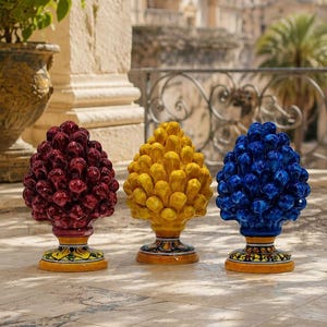 Può includere: Tre sculture in ceramica a forma di pigna nei colori rosso scuro, giallo brillante e blu reale. Ogni pezzo presenta un design testurizzato a scaglie sovrapposte e poggia su una base circolare decorata con motivi dipinti in giallo, verde e nero.