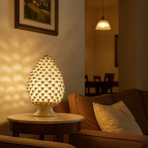 Puede incluir: Lámpara de mesa de cerámica blanca con una pantalla texturizada en forma de piña. La lámpara está sobre una pequeña mesa redonda con tapa de mármol. La luz cálida ilumina un salón con un sofá marrón y una pintura de paisaje enmarcada.