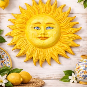 Könnte beinhalten: Handgefertigte gelbe Keramiksonne mit lächelndem Gesicht und blauen Augen. Die Sonne hat detaillierte Strahlen und ist mit "Handcrafted Sicilian Ceramic Sun, Large 44 cm" beschriftet. Der Hintergrund umfasst Zitronen, Blumen und den Text "Ceramica di Caltagirone".
