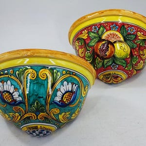 May include: Two ceramic bowls with vibrant hand-painted designs. One bowl features a red background with fruit illustrations, while the other has a teal background with floral patterns. Both have yellow rims and are likely decorative.