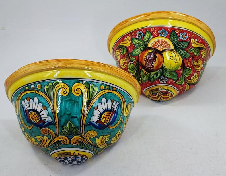 May include: Two ceramic bowls with vibrant hand-painted designs. One bowl features a red background with fruit illustrations, while the other has a teal background with floral patterns. Both have yellow rims and are likely decorative.