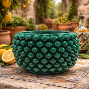 Pode incluir: Um vaso de cerâmica verde esmeralda brilhante com um design texturizado em forma de pinha. O vaso redondo com a parte superior aberta está sobre uma superfície de azulejos, com limões e uma garrafa decorativa no fundo.